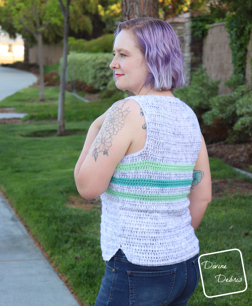 [image description] white woman with purple hair stands facing away from the camera in front of foliage in a white tank top with 3 green stripes, the Hayfield Tank Top free crochet pattern