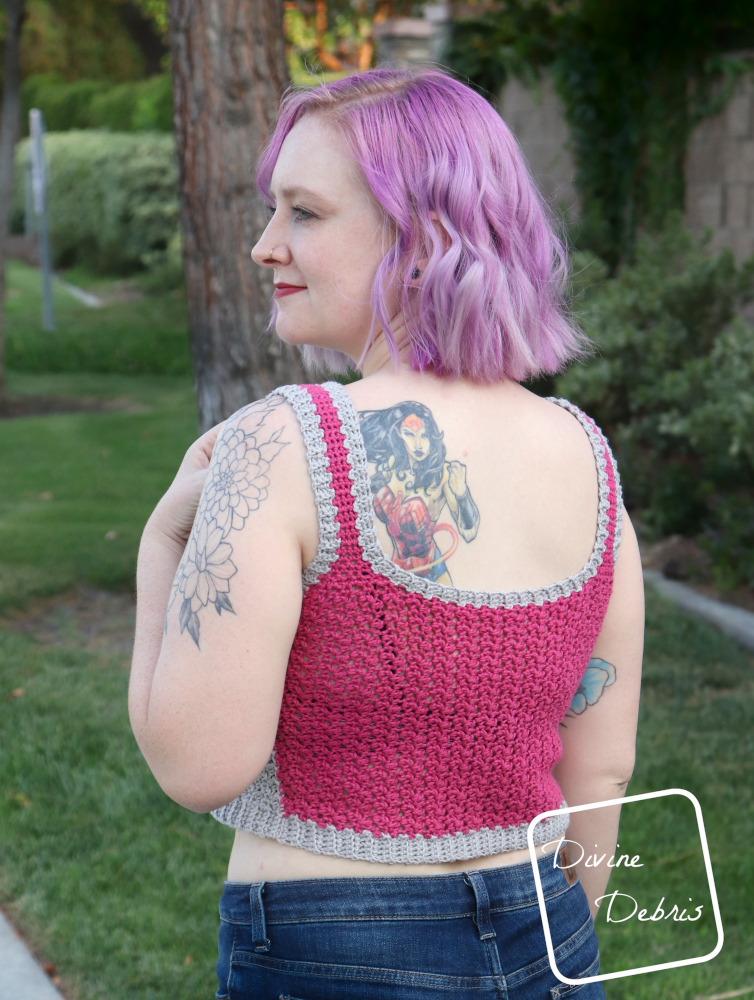 [Image description] A white woman with purple hair stands facing away from the camera in front of a green landscape in the pink and gray Kelsey Tank Top