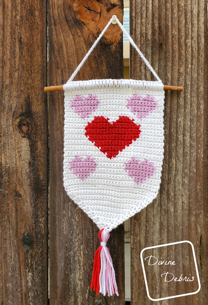 [Image description] Cute Hearts Wall Hanging hangs from a plastic push pin on a wooden fence