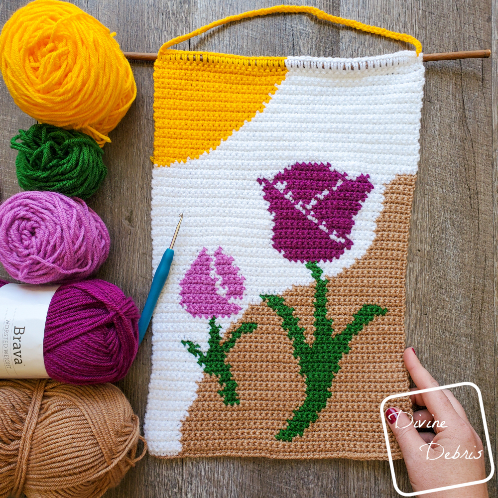 [Image description] Top down view of the Cute Tulips Wall Hanging on a wood grain background, skeins of yarn and a crochet hook on the left of the wall hanging and a white woman's hand holding the unfinished bottom left corner.