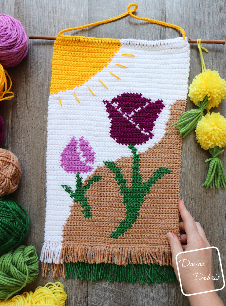 [Image description] The Cute Tulips Wall Hanging laying flat on a wood grain background, with skeins of yarn on the left and a white woman's hand holds the bottom left corner.