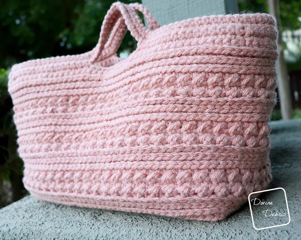 [Image description] A low angle photo of the soft pink Diana Basket on grey piece of concrete in front of a grey post and plants to the left.