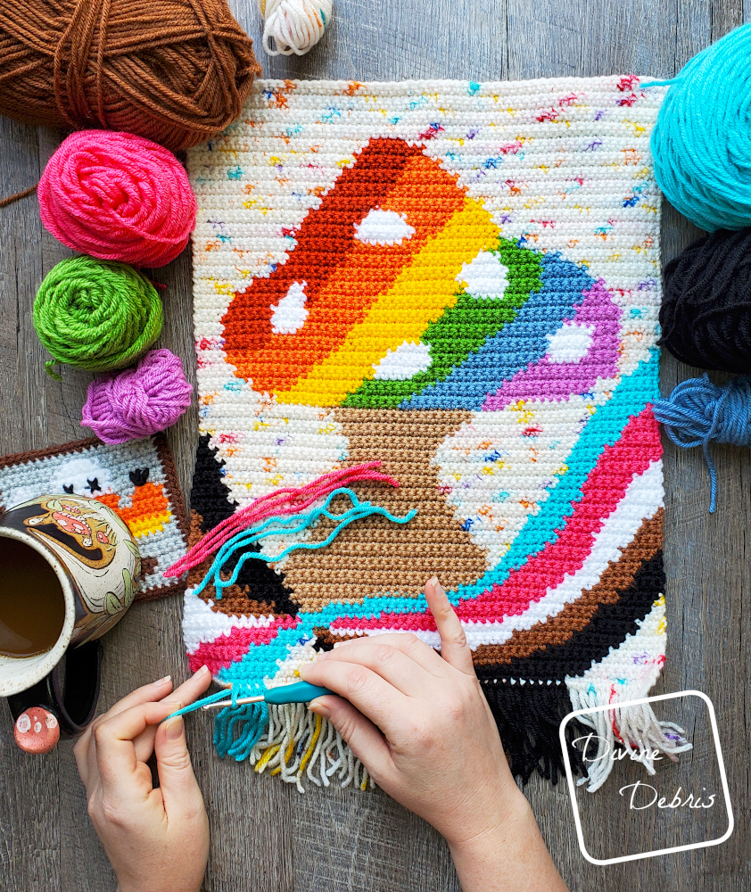 [Image description] Top down view of the 'Shroom For Everyone Wall Hanging on a wood grain background with cakes of yarn along the left and right sides, with a white woman's hands attaching fringe to the bottom.