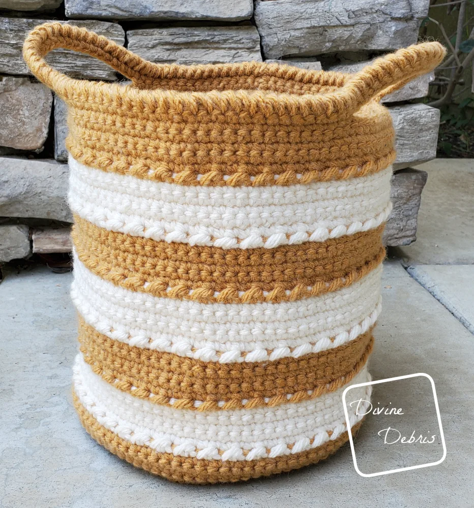 [Image description] Yellow and white striped Claire Basket sits on a black shelf against a stone background on a cement floor