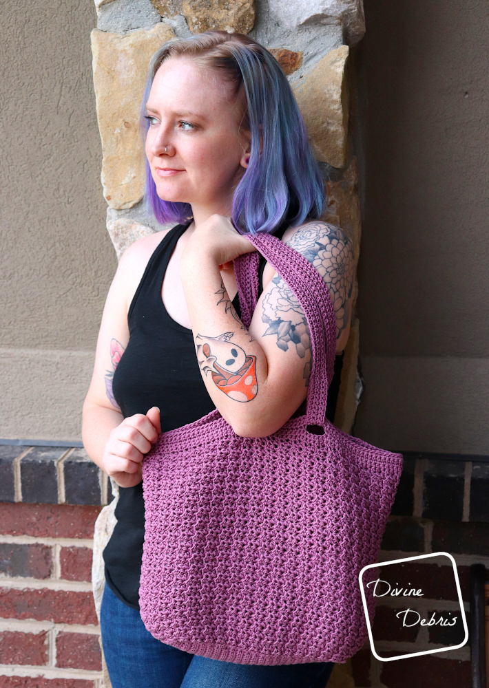 [Image description] A white woman with blue hair stands against a brick wall looking to her right, holding the purple Lucinda Bag.