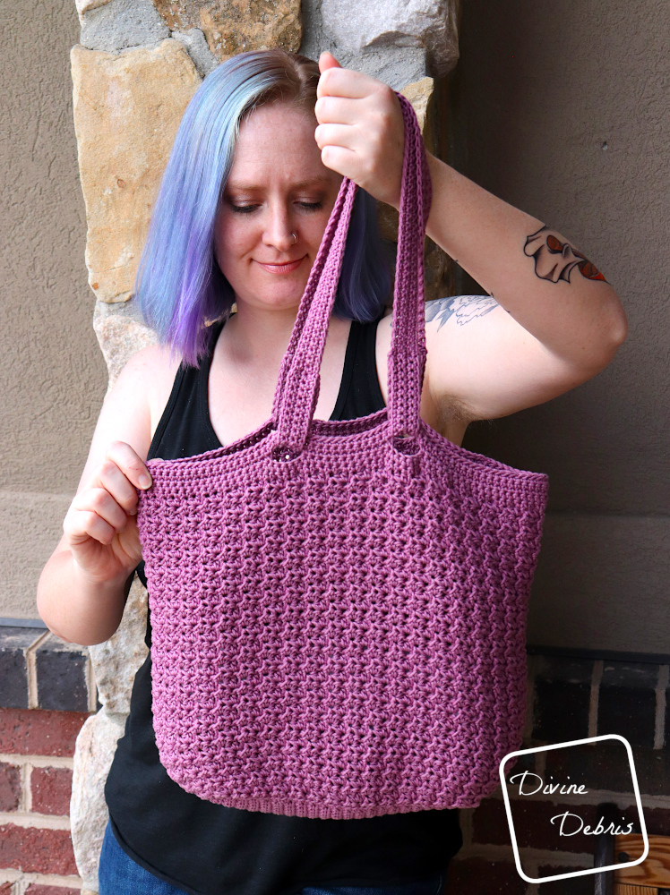 [Image description] a white woman with blue hair stands against a brick wall holding the Lucinda Bag up toward the camera.
