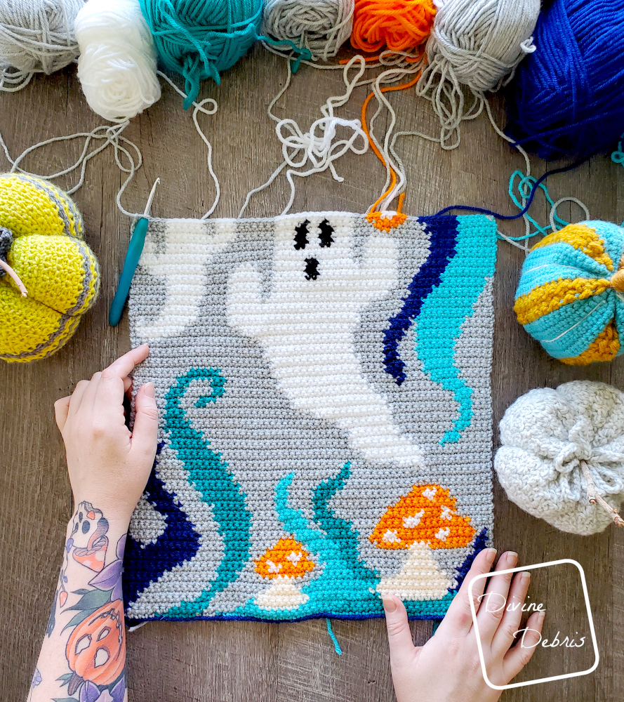 [Image description] a half finished Ghosts of the Blue Lagoon Wall Hanging laying on a wood grain background surrounded by skeins of yarn above it and a white woman's hands on the bottom.