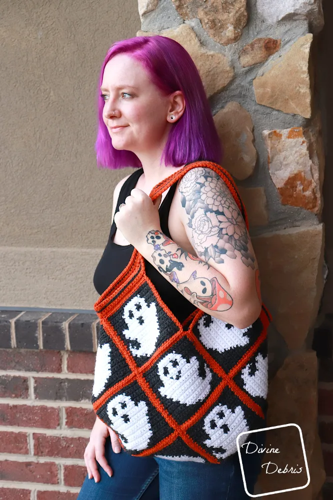 [Image description] A white woman with purple hair stands against a brick wall looking to her right, holding the Glenda Ghost Bag.